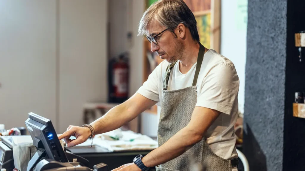 store-owner-wearing-eyeglasses-using-cash-register-at-checkout-in-organic-market-model-released-symb.webp