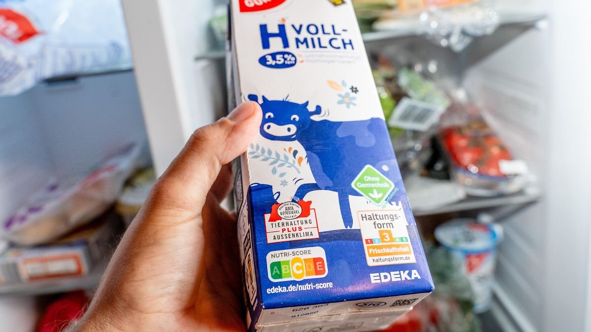 Eine-Hand-haelt-eine-H-Vollmilch-vor-dem-geoeffneten-Kuehlschrank.jpg