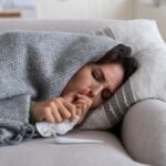Young-woman-lying-under-blanket-on-sofa-coughing-and-sneezing-using-paper-tissue-to-blow-her-nose-Un.jpeg