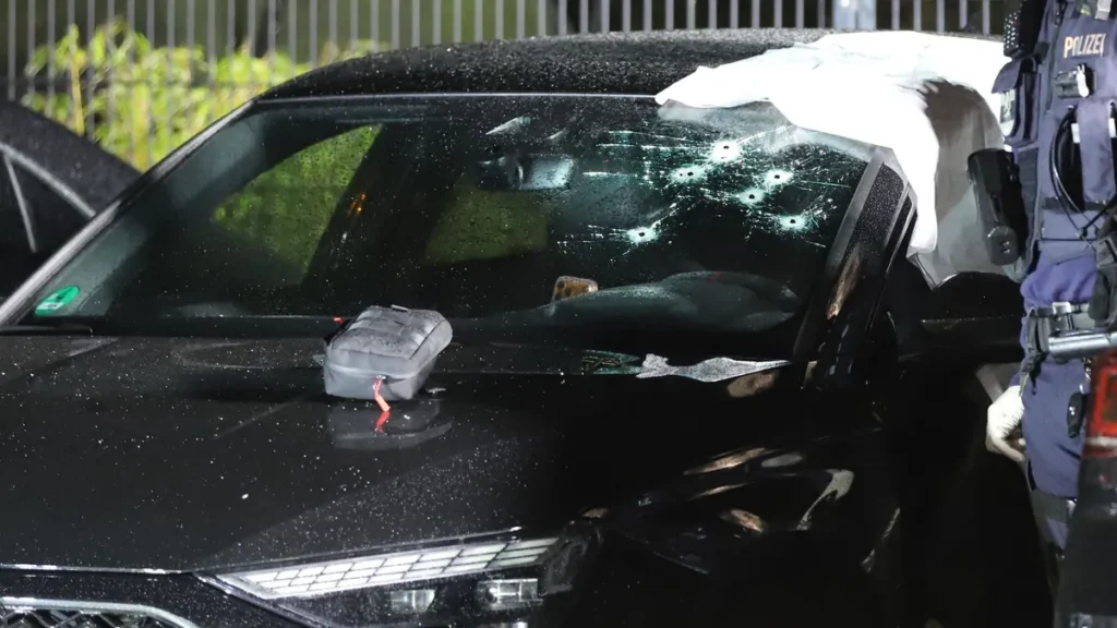 einsatzkraefte-der-polizei-stehen-an-einer-durch-schuesse-getroffenen-windschutzscheibe-von-einem-pk.webp
