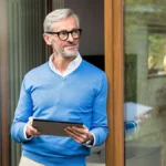 smiling-senior-man-with-grey-hair-standing-in-front-of-his-modern-design-home-holding-tablet-model-r.webp