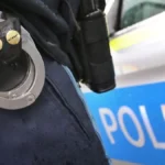 ein-polizist-mit-handschellen-und-einer-pistole-am-guertel-steht-vor-einem-streifenwagen-symbolbild.webp