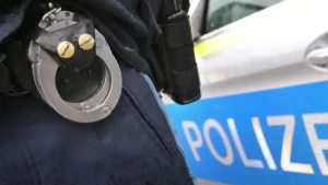 ein-polizist-mit-handschellen-und-einer-pistole-am-guertel-steht-vor-einem-streifenwagen-symbolbild.webp