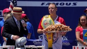 Joey-Chestnut-a-multi-time-winner-holds-a-plate-of-hot-dogs-with-the-Mustard-Belt-over-his-shoulder.jpeg